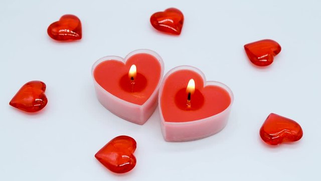 The blurred frame becomes clear: Two bright red heart-shaped candles burn on a white background with decoration of small glass hearts. The Candles burn for a while, at the end they are blown out.