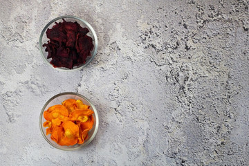 Dried carrot and beet chips in a glass bowl on a gray concrete background. Organic natural food. Top view, flat lay.