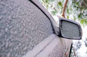 Snow covered car on a pine background, snow covered side window of a car and side view mirror