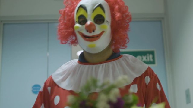 A clown walking down a hallway holding a bouquet of flowers as the camera dollies backwards