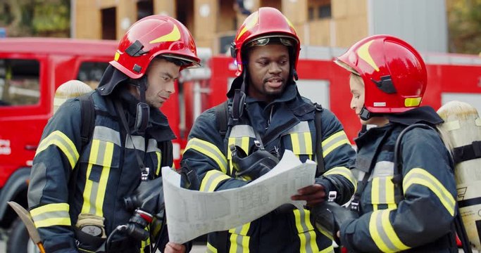 Caucasian pretty woman and wo multiethnic men fire fighters co-workers standing outdoors at the fire truck with a big plan or map in hands and talking while deciding how to fight with fire.