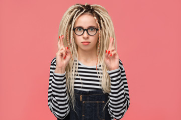 Fototapeta premium Young girl with long dreads standing over pink background holding fingers crossed for good luck