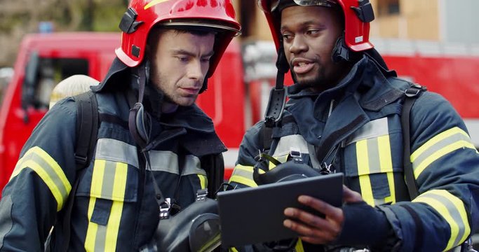 Mixed-races firemen co-workers in helmets and special costumes standing at teh fire truck outside and discussing something with a tablet device in hands.