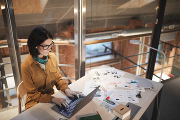 Serious fashion designer sitting at her workplace among sketches and typing on laptop computer at...