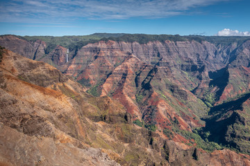 Fototapeta na wymiar Waimea Canyon, Kauai, Hawaii, USA. - January 16, 2020: Wide landscape with waterfall in distance shows red rocks partly green in crevasses, green cover on top, under blue sky.