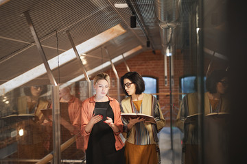 Young businesswoman using digital tablet and talking to her colleague with note pad while they walking along the office corridor