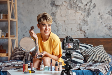 Make-up Blogger. Young woman sitting at stylish apartment recording video on camera applying rouge...