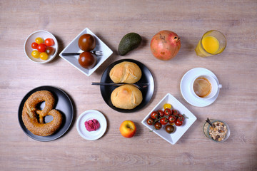 Pretzel, sweet tea-cake, marmelade, avocado, tomatoes, a pomegranate and a glass of orange juice, symetrically arranged on a wooden table on white and black tableware