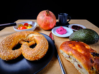 Breakfast table with pretzel, sweet tea-cake, marmelade, avocado, tomatoes, a pomegranate, a glass of orange juice, on a wooden table and in front of a dark background