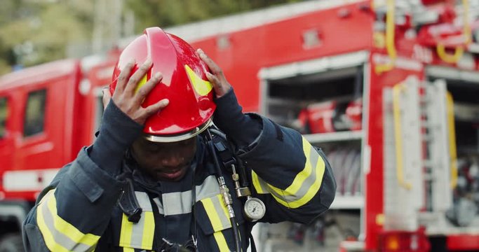 Portrait shot of the African American joyful fire fighter taking off helmet and smiling to the camera at the big red truck after fire.