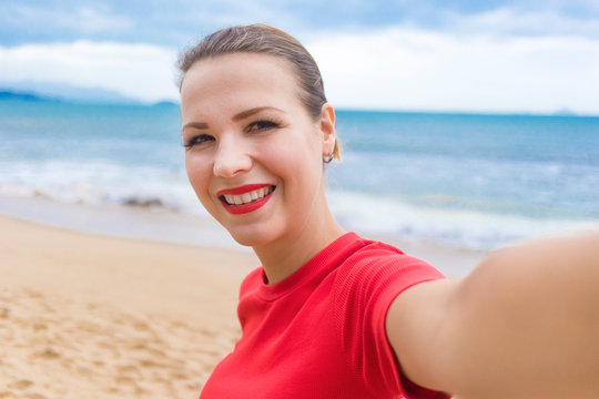 Happy positive cheerful girl, young woman doing, taking selfie, photo, picture of herself on mobile smartphone camera on vacation on the beach, sea background, smiling, laughing, having fun outdoors