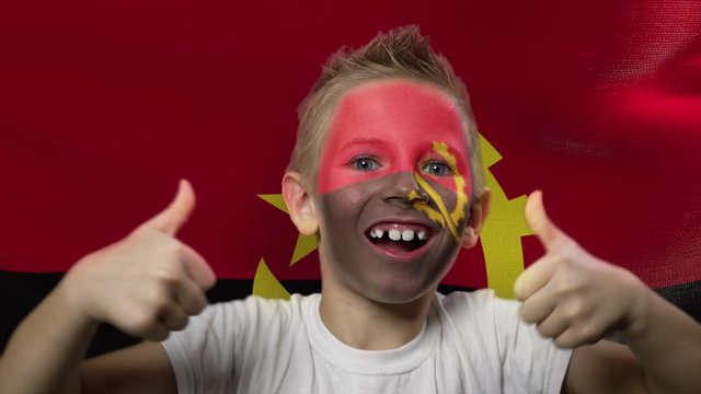 Joyful fan on the background of the flag of Angola. Happy boy with painted face in national colors. The young fan rejoices in the victory of his beloved team. Success. Victory. Triumph.