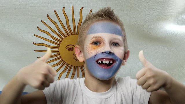 Joyful fan on the background of the flag of Argentina. Happy boy with painted face in national colors. The young fan rejoices in the victory of his beloved team. Success. Victory. Triumph.