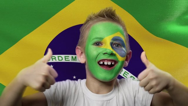 Joyful fan on the background of the flag of Brazil. Happy boy with painted face in national colors. The young fan rejoices in the victory of his beloved team. Success. Victory. Triumph.