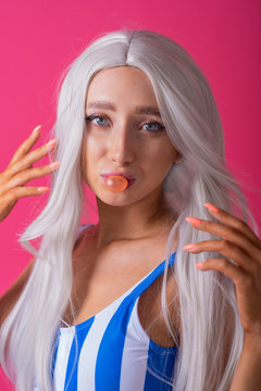 Portrait of a slender woman in a blonde wig posing on a pink background. Cute girl with blue eyes in striped swimsuit chewing gum and blowing bubbles.
