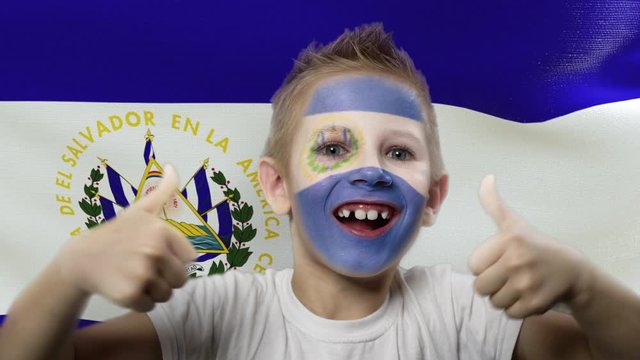 Joyful fan on the background of the flag of El Salvador. Happy boy with painted face in national colors. The young fan rejoices in the victory of his beloved team. Success. Victory. Triumph.