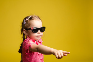 Stock photo of cute little girl with hair accessories wearing sunglasses and pink dress showing the direction with her index finger. Isolate on yellow background.