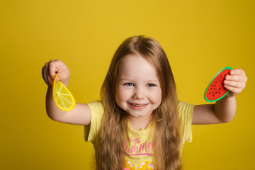 Front view of happy girl standing on yellow isolated background and closing eyes with toys. Cheerful longhaired child laughing and playing hide and seek. Concept of game and happiness.