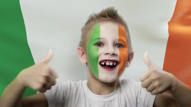 Joyful fan on the background of the flag of the Ireland. Happy boy with painted face in national colors. The young fan rejoices in the victory of his beloved team. Success. Victory. Triumph.