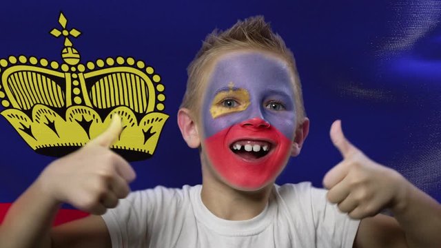 Joyful fan on the background of the flag of Liechtenstein. Happy boy with painted face in national colors. The young fan rejoices in the victory of his beloved team. Success. Victory. Triumph.