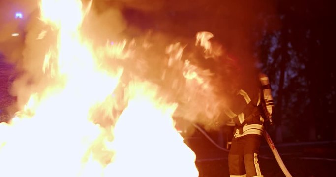 Fire fighter in the equipped costume, helmet and gas mask extinguish the fire with water late at night. Outdoors.