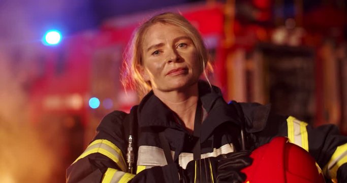 Portrait of the Caucasian tired female firefighter with the helmet in hands looking to the camera with a big fire truck on the background at night. Outdoor.