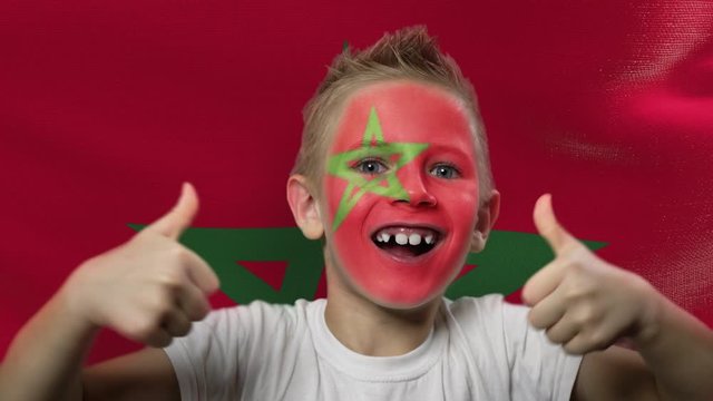 Joyful fan on the background of the flag of Morocco. Happy boy with painted face in national colors. The young fan rejoices in the victory of his beloved team. Success. Victory. Triumph.