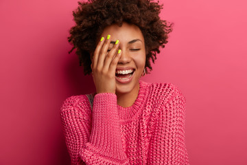 Emotions, lifestyle concept. Overjoyed dark skinned woman laughs with closed eyes, hears good joke, covers face with palm, has nice conversation, chuckles and shows white teeth, wears knitted sweater