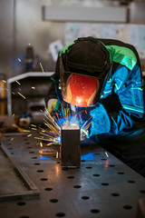 Worker welding two pieces of metal square tube to form a 90 degree angle on a work bench in a factory workshop - obrazy, fototapety, plakaty