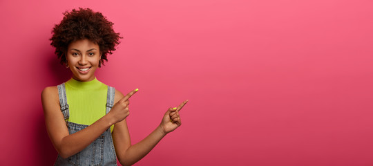 Studio shot of smiling curly woman wears bright vest and dungarees, points both index fingers at upper right corner, demonstrates good copy space for your promotional content, isolated on pink wall - obrazy, fototapety, plakaty