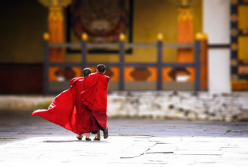 Konigreich Bhutan, Asien, Himalaya. Zwei junge Novizen in Innenhof eines Kloster laufen umarmt zum Tempel. Ihre roten Gewänder leuchten in der Sonne und wehen im Wind.