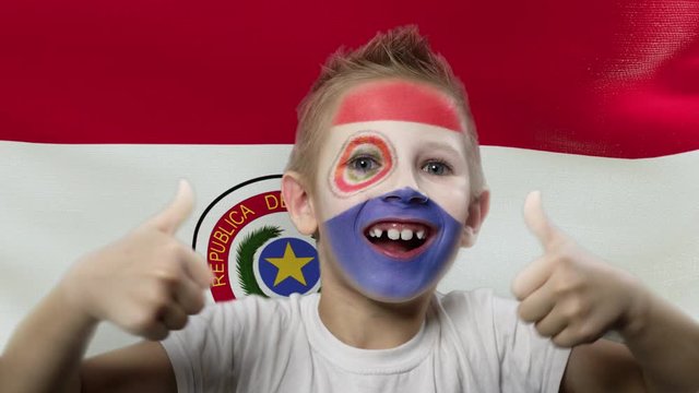 Joyful fan on the background of the flag of Paraguay. Happy boy with painted face in national colors. The young fan rejoices in the victory of his beloved team. Success. Victory. Triumph.