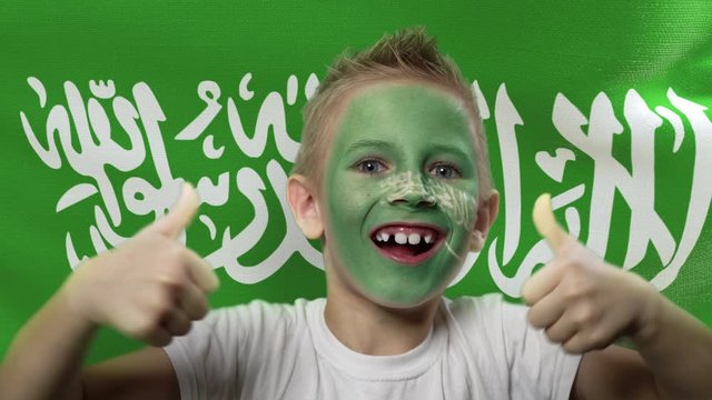 Joyful fan on the background of the flag of Saudi Arabia. Happy boy with painted face in national colors. The young fan rejoices in the victory of his beloved team. Success. Victory. Triumph.