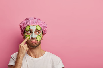Puzzled man points at face, looks aside, claims on having blackheads, applies cucumbers and clay mask, wears bath cap, purses lower lip, isolated over pink wall, copy space aside. Beauty treatment