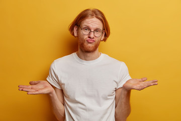 Hesitant red head male youngster spreads palms, shrugs shoulders with bewilderment, pouts lips and feels confused, chooses something, wears optical glasses and t shirt, faces difficult choice