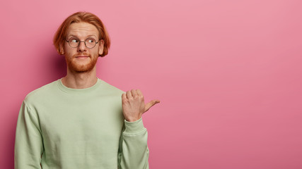Bearded red head adult man points with thumb to right side on copy space, shows advertisement or...