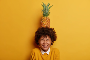 Indoor shot of overjoyed Afro American woman carries pineapple on head, closes eyes and smiles broadly, has fun with tropical fruit, isolated over yellow bright background. People, emotions concept