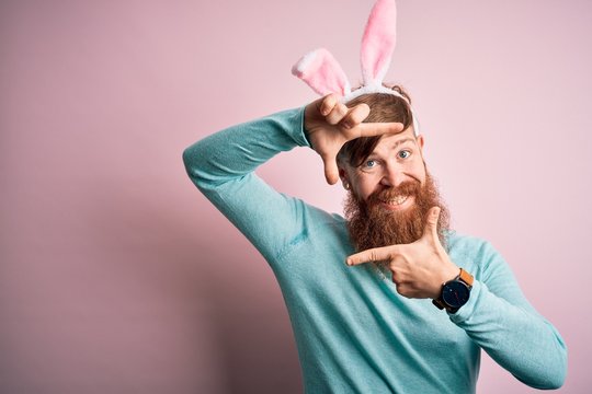 Hipster Irish man with beard wearing easter rabbit ears over isolated pink background smiling making frame with hands and fingers with happy face. Creativity and photography concept.