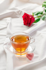 Beautiful romantic morning with cup of tea, heart shaped candy and red rose on white bed