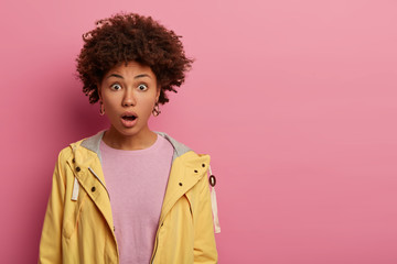 Obraz na płótnie Canvas Cute curly haired young woman gasps from fear, keeps jaw dropped, afraids of something, stands with amazement and concern, sees unbelievable thing, isolated on pink wall, copy space for text