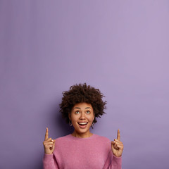 Cheerful smiling ethnic woman points above, advertises cool object, says check out promo, has glad...