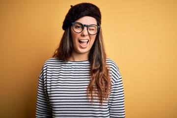Young beautiful brunette woman wearing french beret and glasses over yellow background winking...