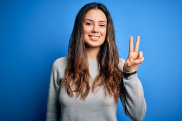 Young beautiful brunette woman wearing casual sweater standing over blue background smiling with happy face winking at the camera doing victory sign. Number two.