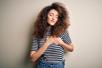 Young beautiful woman with curly hair and piercing wearing casual striped t-shirt smiling with hands on chest with closed eyes and grateful gesture on face. Health concept.