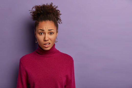Human facial expressions concept. Displeased Afro woman smirks face and purses lips, reacts on hearing something unpleasant, wears casual jumper, models over purple wall with blank space aside