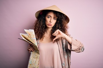 Young beautiful tourist woman with curly hair and piercing wearing asian hat holding city map with...
