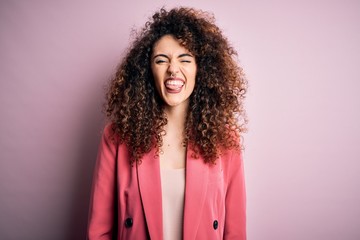Young beautiful businesswoman with curly hair and piercing wearing elegant jacket sticking tongue...