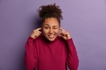 Relieved glad ethnic woman plugs ears, winks eyes, hears no sound or noise, smiles broadly, attends very loud concert, dressed casually, has curly combed hair. Human emotions and reaction concept