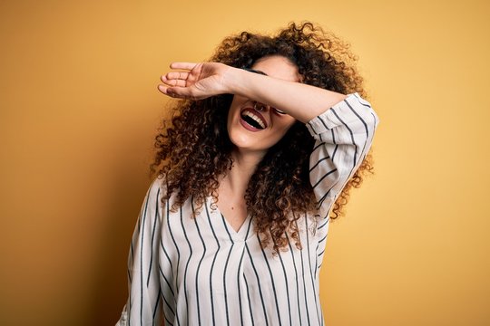 Young beautiful woman with curly hair and piercing wearing striped shirt and glasses covering eyes with arm smiling cheerful and funny. Blind concept.