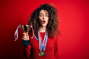 Young beautiful successful woman with curly hair and piercing holding trophy wearing medals scared in shock with a surprise face, afraid and excited with fear expression